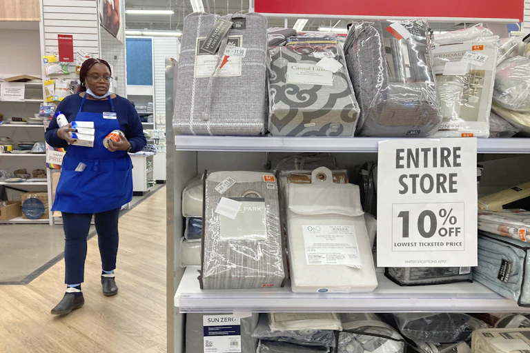 a black woman wearing a blue bed bath and beyond apron holds some products as she walks through aisles at a store, where a sign near bedding materials reads entire store 10% off