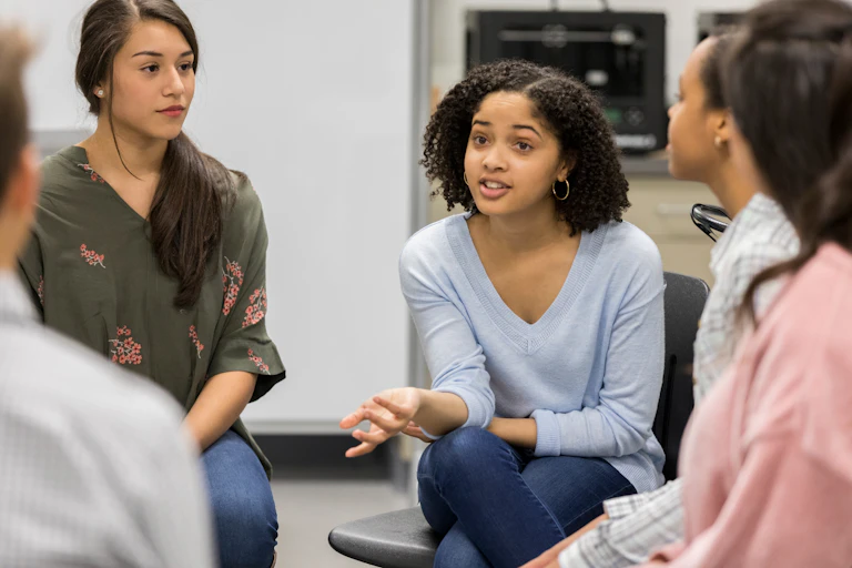 Teenage girls sit in chairs in a circle talking to one another. It's a support group.