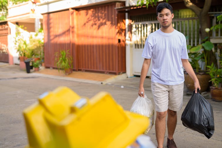 An Asian man carrying two trash bags towards a dumpster