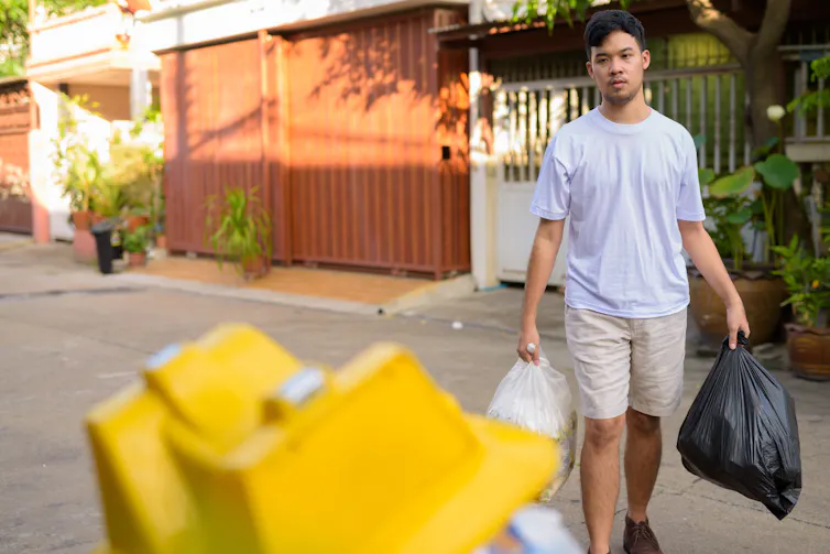 An Asian man carrying two trash bags towards a dumpster