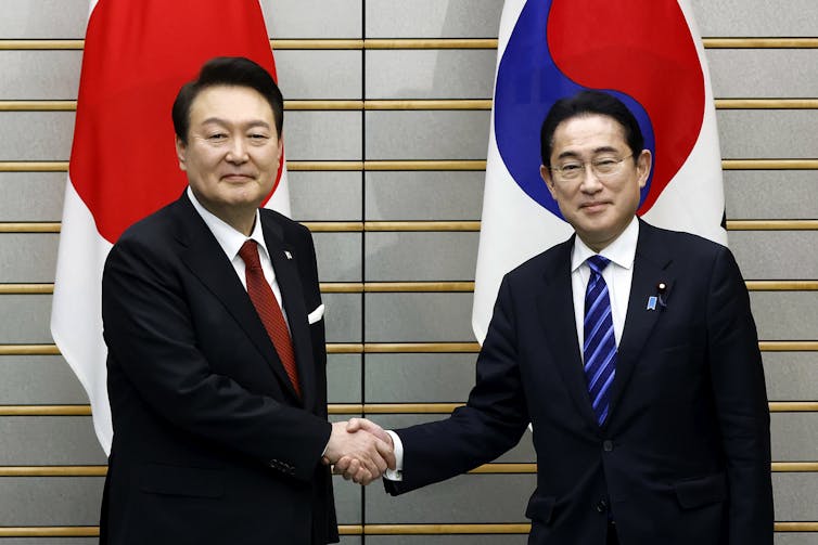 Two men shake hands in front of a South Korean and Japanese flag.
