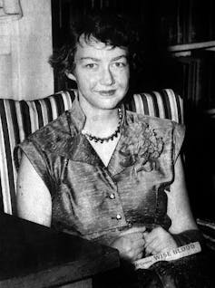 A woman with short hair, a necklace, and a short-sleeve dress smiles guardedly in a black and white photograph.