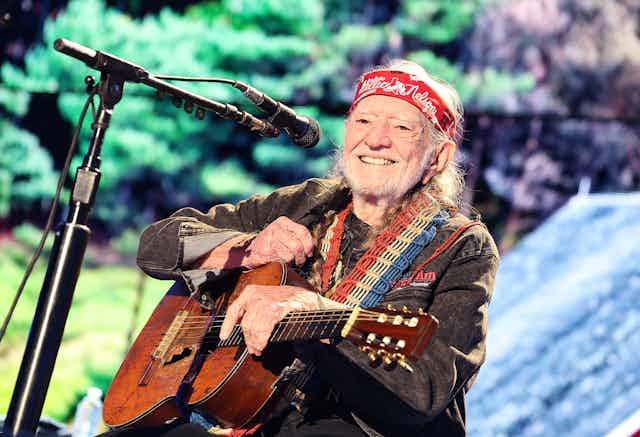 a bearded old man wearing a bandanna on his forehead sits on a stage in front of a microphone holding a beat up acoustic guitar