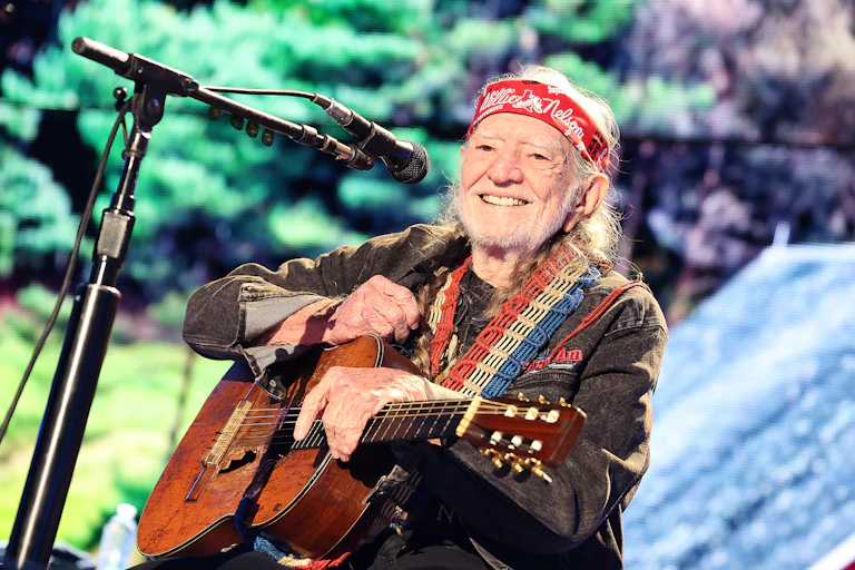 a bearded old man wearing a bandanna on his forehead sits on a stage in front of a microphone holding a beat up acoustic guitar
