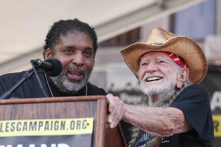 two bearded men, one Black and middle-aged and the other white and elderly and wearing a straw hat, stand together at a podium on a stage