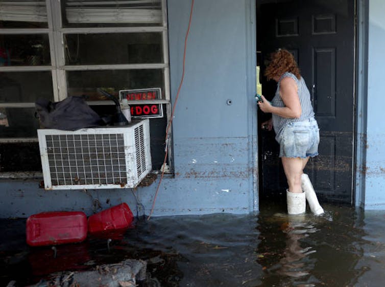 A woman standing in floodwater in boots looks in the door of a flooded home. A sign reading 'beware of dog' is in the window. Floodwater is up to her shins.