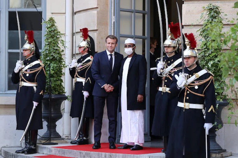 Un homme en costume-cravate gris serre la main d'un autre homme en robe et casquette blanches.