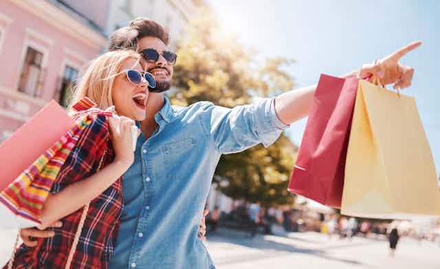 A young man and woman outdoors with their arms around each others waists smiles excitedly and looks at something in the distance while the man points, both are wearing trendy clothing and carrying multiple shopping bags, with sunglasses
