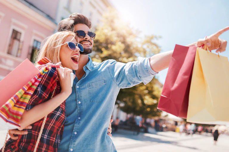 A young man and woman outdoors with their arms around each others waists smiles excitedly and looks at something in the distance while the man points, both are wearing trendy clothing and carrying multiple shopping bags, with sunglasses