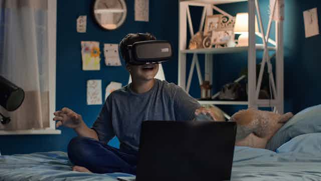Boy in bedroom with laptop using VR headset
