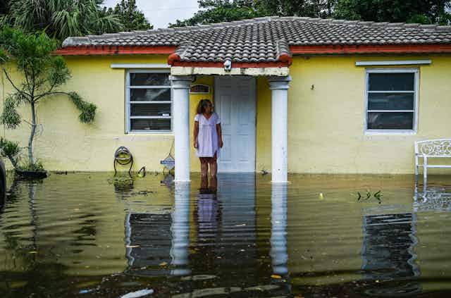 A woman stands in front of a small single-story house, shin-deep in flood water.