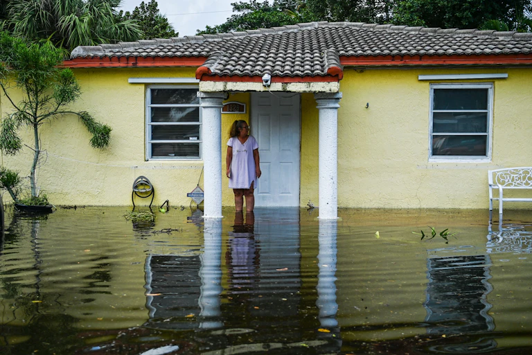 A woman stands in front of a small single-story house, shin-deep in flood water.
