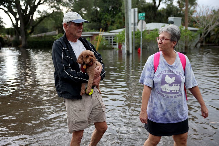 An older man carries a small fuzzy dog while walking through floodwater with a woman waring a backpack.