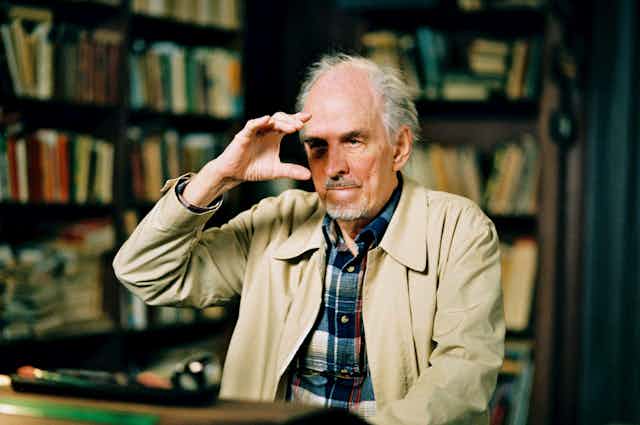 Ingmar bergman in 2003 wearing a plaid shirt and beige jacket. He holds his hand to his face as if holding a camera. 