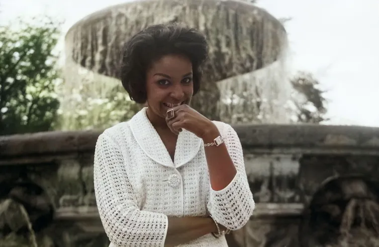 Singer Mona Baptiste in a white cardigan posing in front of a fountain.