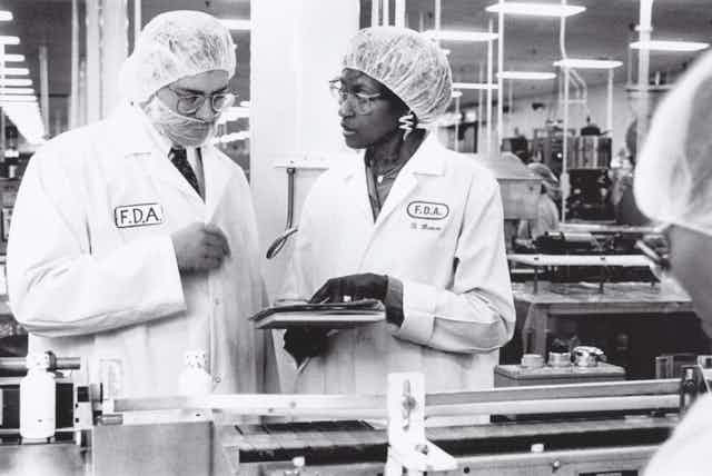 Two people wearing hair nets and FDA white coat inspect batch records at a drug assembly line