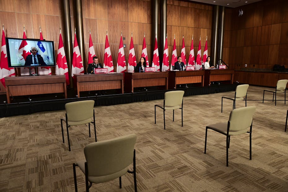 People sit at a long table in front of a row of Canadian flags while a man appears on screen. Empty chairs several feet apart sit in front of the long table.