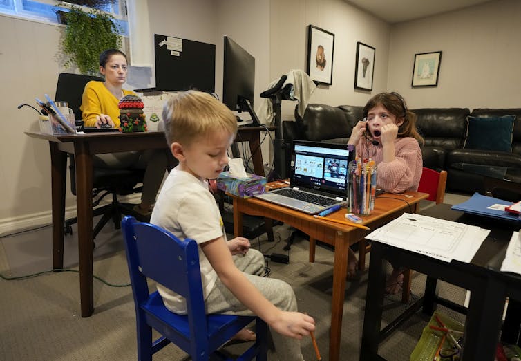 Two children sit at laptops while their mother sits at a desk looking at her own laptop.