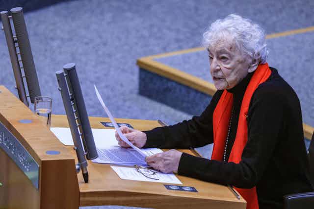 An elderly woman in a black top and bright red scarf speaks into microphones at a lecturn, holding up a paper she is evidently reading from.