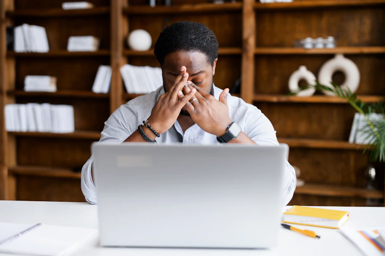 A man sitting at a laptop and wearing a collared shirt covers his face with his hands, clearly frustrated