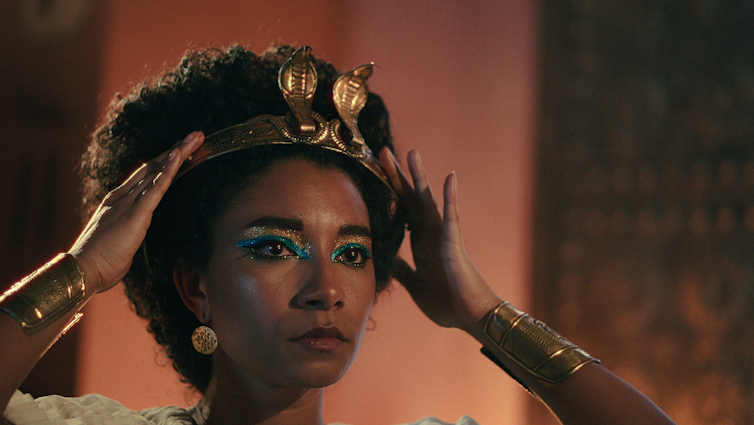 Cleopatra lifts a crown to her head, her eyes lined with blue kohl. She is a black woman.