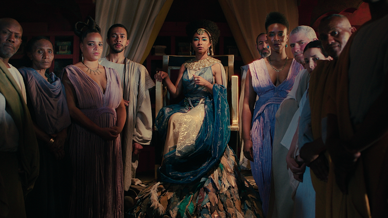 The cast stand around Cleopatra's throne in robes.