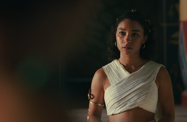 Cleopatra in a white cross over top with gold jewellery. She is a black woman.
