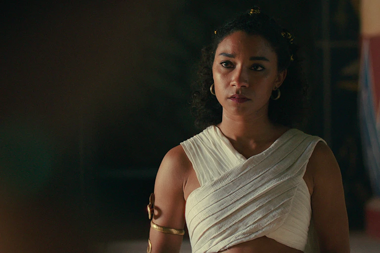 Cleopatra in a white cross over top with gold jewellery. She is a black woman.