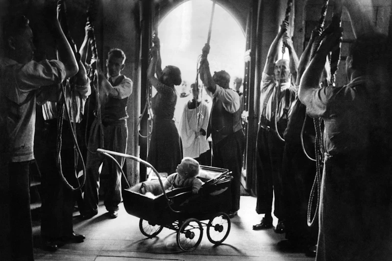 A baby in a buggy looks up at adults pulling on bell ropes.