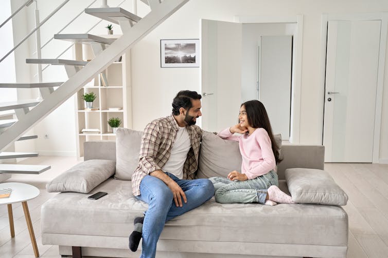 Ayah dan anak perempuan duduk di sofa sambil berbincang dan tersenyum