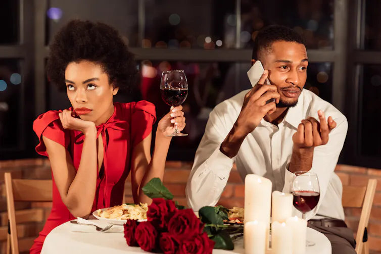Man talking on his phone sat next to a woman holding a glass of wine looking annoyed.