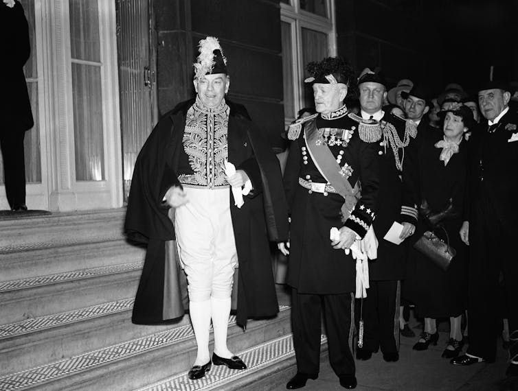 A man in a fancy hat and military uniform stands on the steps of a hotel.