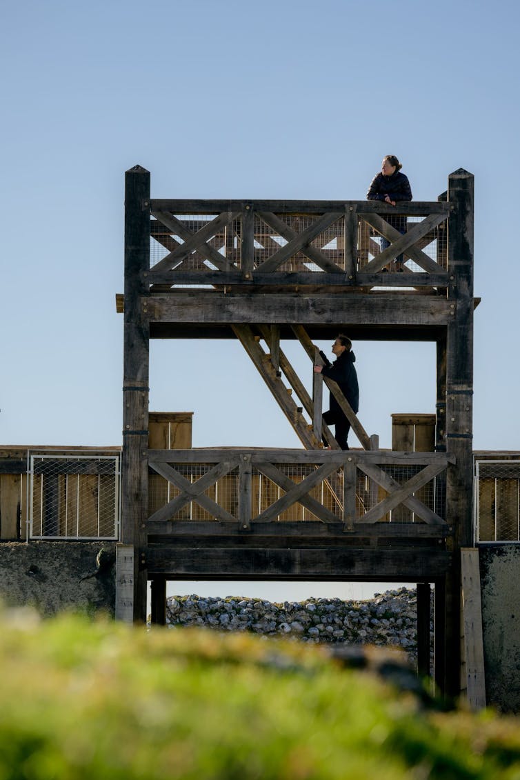 Two storey wooden platform above a ditch with people climbing stairs between levels.