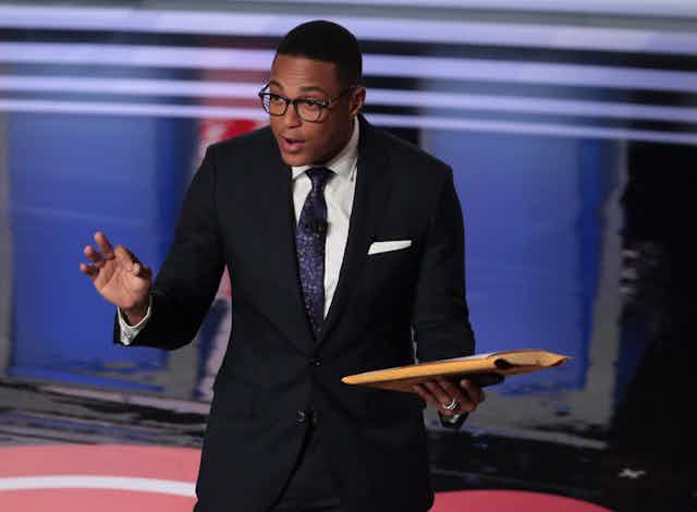 A black man wearing eyeglasses and dressed in a business suit holds a book while standing on a stage. 