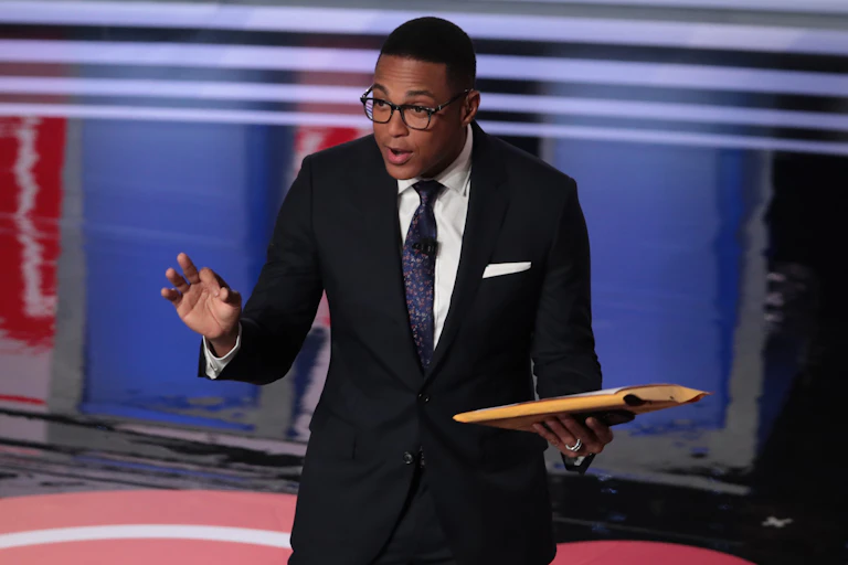 A black man wearing eyeglasses and dressed in a business suit holds a book while standing on a stage.