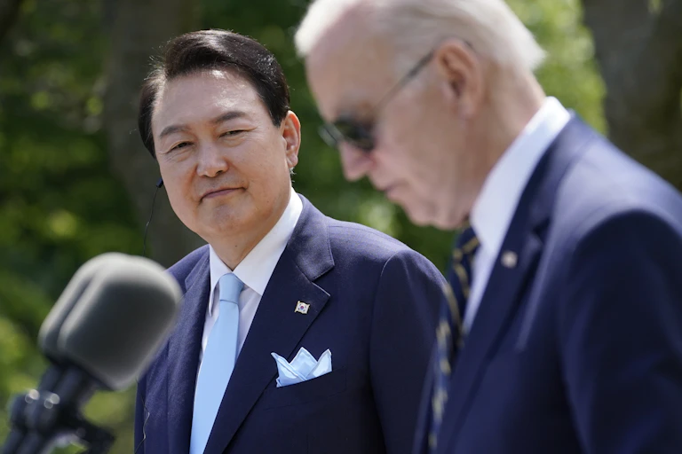 A smiling man in a blue suit looks across to another elderly man in sunglasses