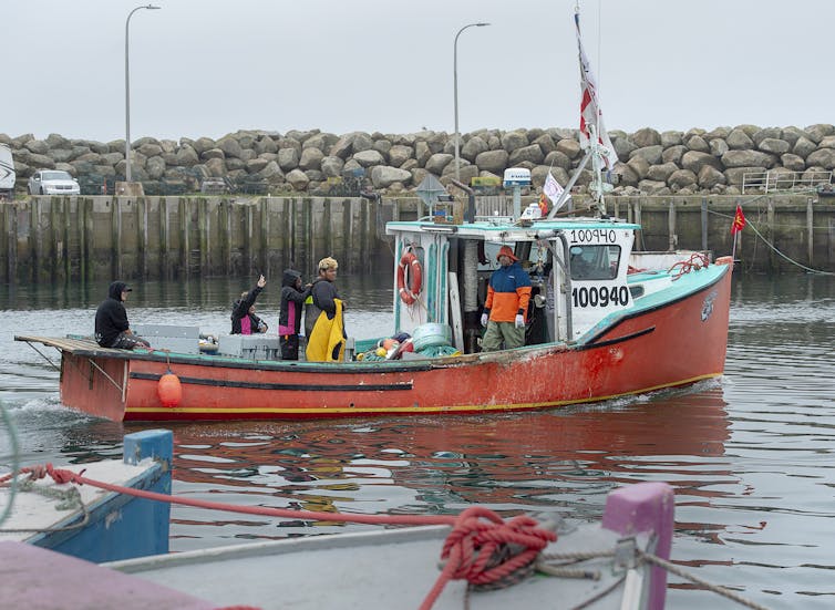 People stand on the deck of a small fishing boat that is floating in the harbour of a body of water.