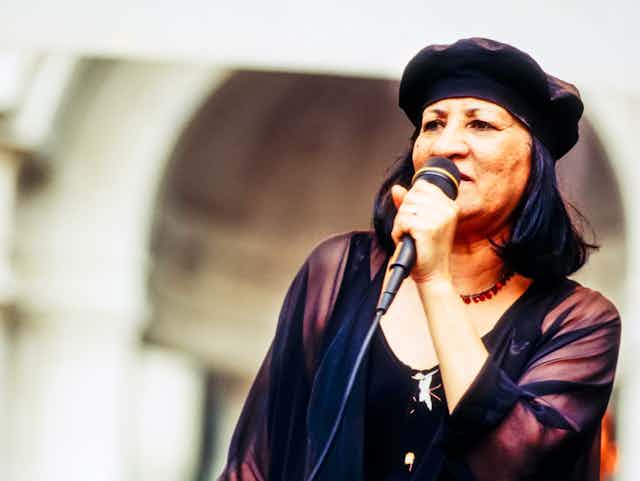 A woman in a black beret and black blouse sings into a microphone in a sepia toned image.