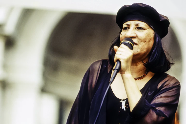 A woman in a black beret and black blouse sings into a microphone in a sepia toned image.
