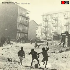 An album cover featuring a sepia toned photo of young boys playing soccer in a street with high-rise housing and shacks.