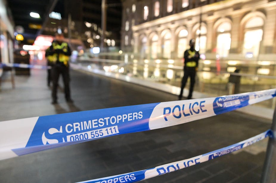 A close up of a blue and white police tape with the word 'Crimestoppers 0800555111' on it. In the background, several police officers stand cross armed. 