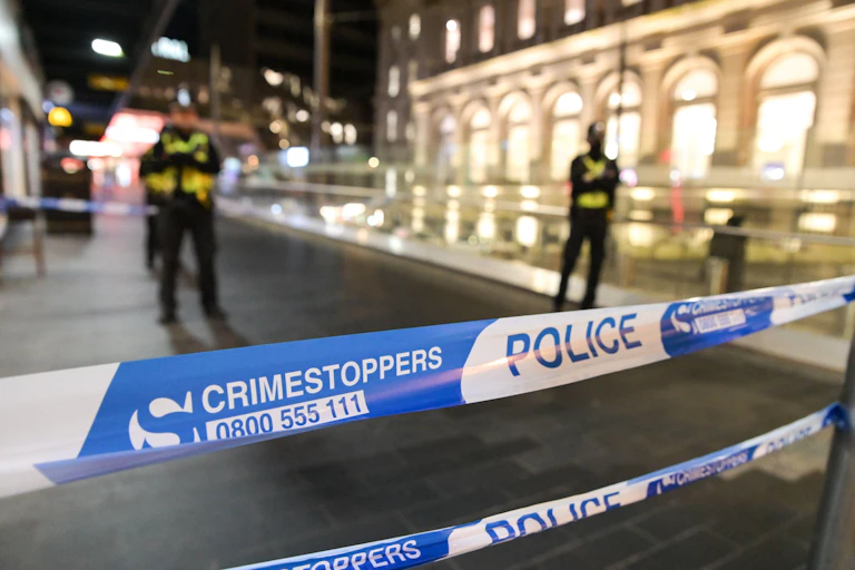 A close up of a blue and white police tape with the word 'Crimestoppers 0800555111' on it. In the background, several police officers stand cross armed.