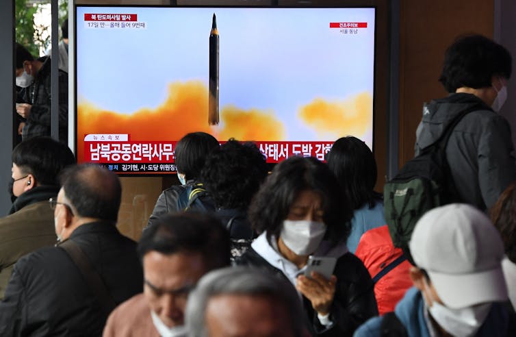 People walk in front of a TV screen showing a missile taking off.
