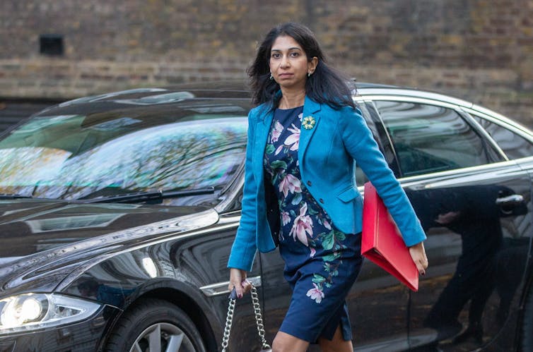 Home Secretary Suella Braverman walking outdoors in front of a black car, holding a red minister's folder under her arm