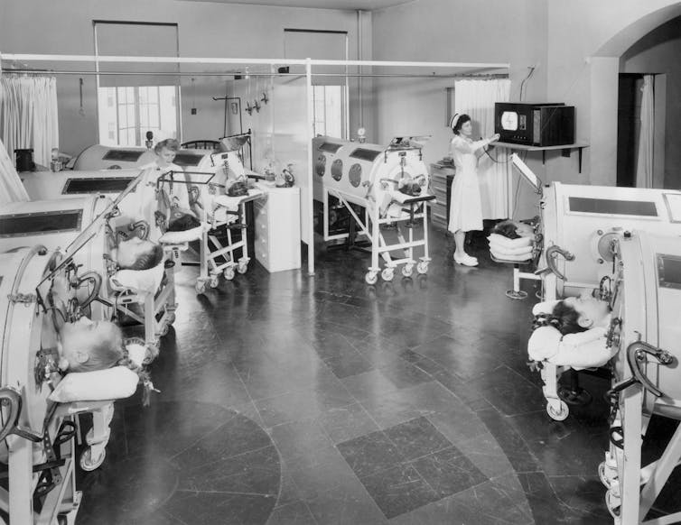 semicircle of patients in iron lungs use mirrors to watch a TV