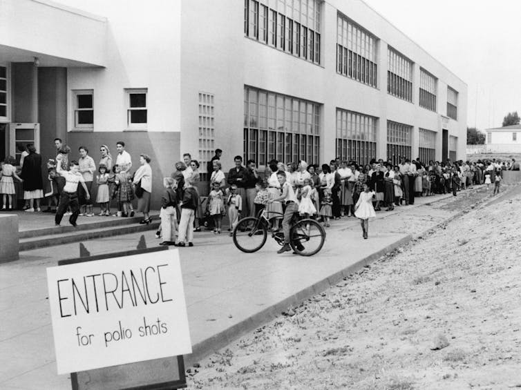 families in line outside a school with a sign 'Entrance for polio shots' in 1955