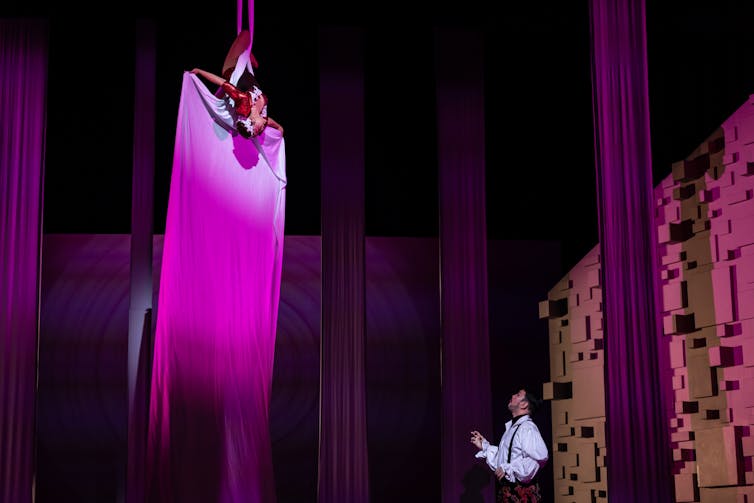 Acrobat hanging upside down, singing to man standing on stage