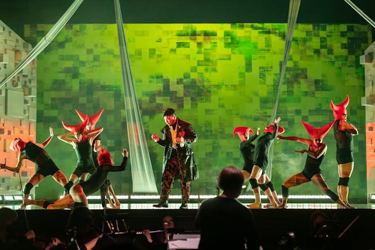 A figure seen clenching his fists on a stage surrounded by dancing figures wearing double-horned hats.