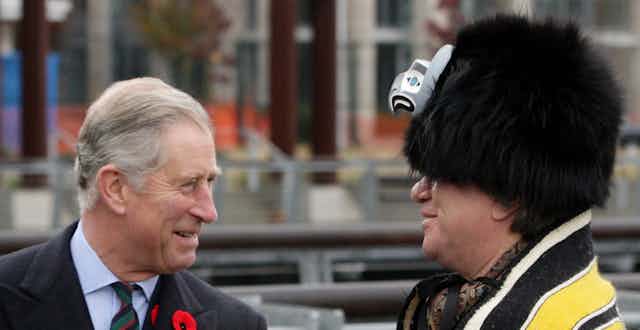 A man in suit wearing poppy speaks with a man in West Coast Indigenous regalia including a hat with an eagle figure on the front and a yellow blanket. 