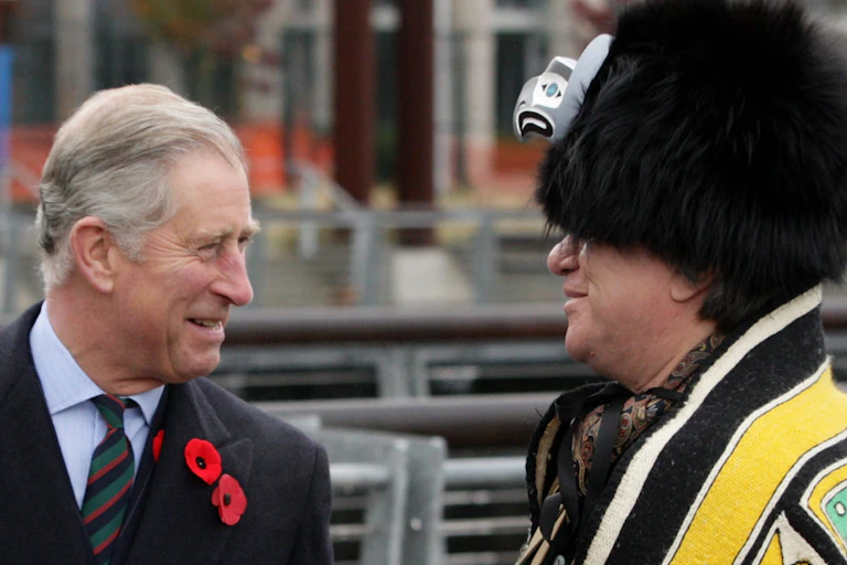 A man in suit wearing poppy speaks with a man in West Coast Indigenous regalia including a hat with an eagle figure on the front and a yellow blanket.